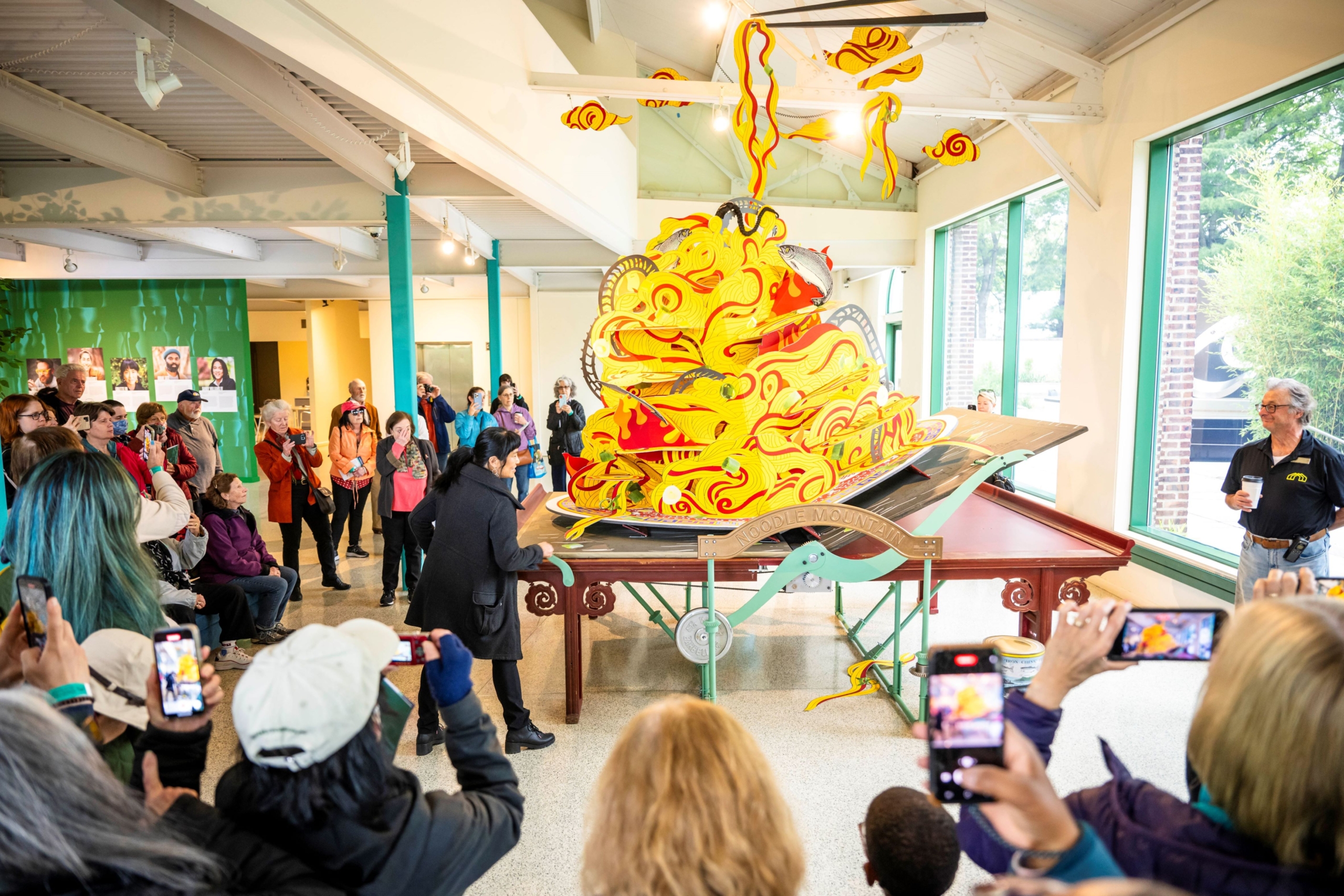 Artist Colette Fu turns the handle to begin to close her pop-up book sculpture, Noodle Mountain, in front of a crowd of people.