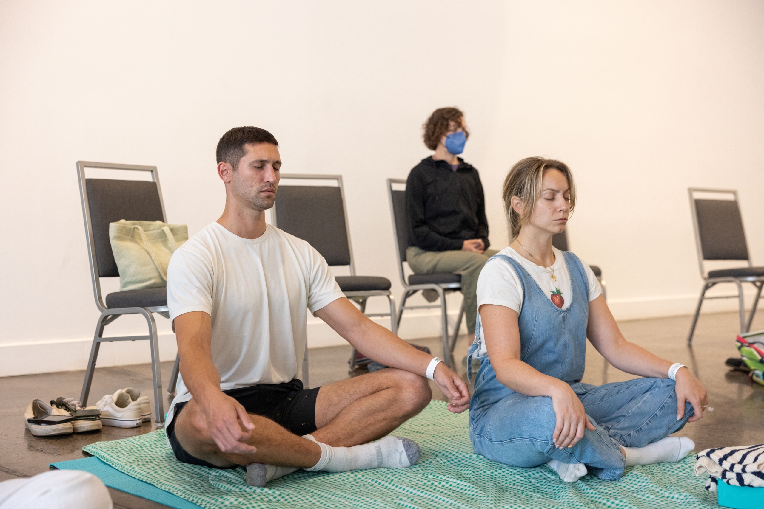 A man and a woman seated on yoga mats with their legs crossed. A second woman in a blue mask sits in a chair behind them.